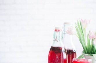 Duas garrafas com kombucha, de coloração avermelhada e flores decorando a mesa. Foto: Brooke Lark ia Unsplash